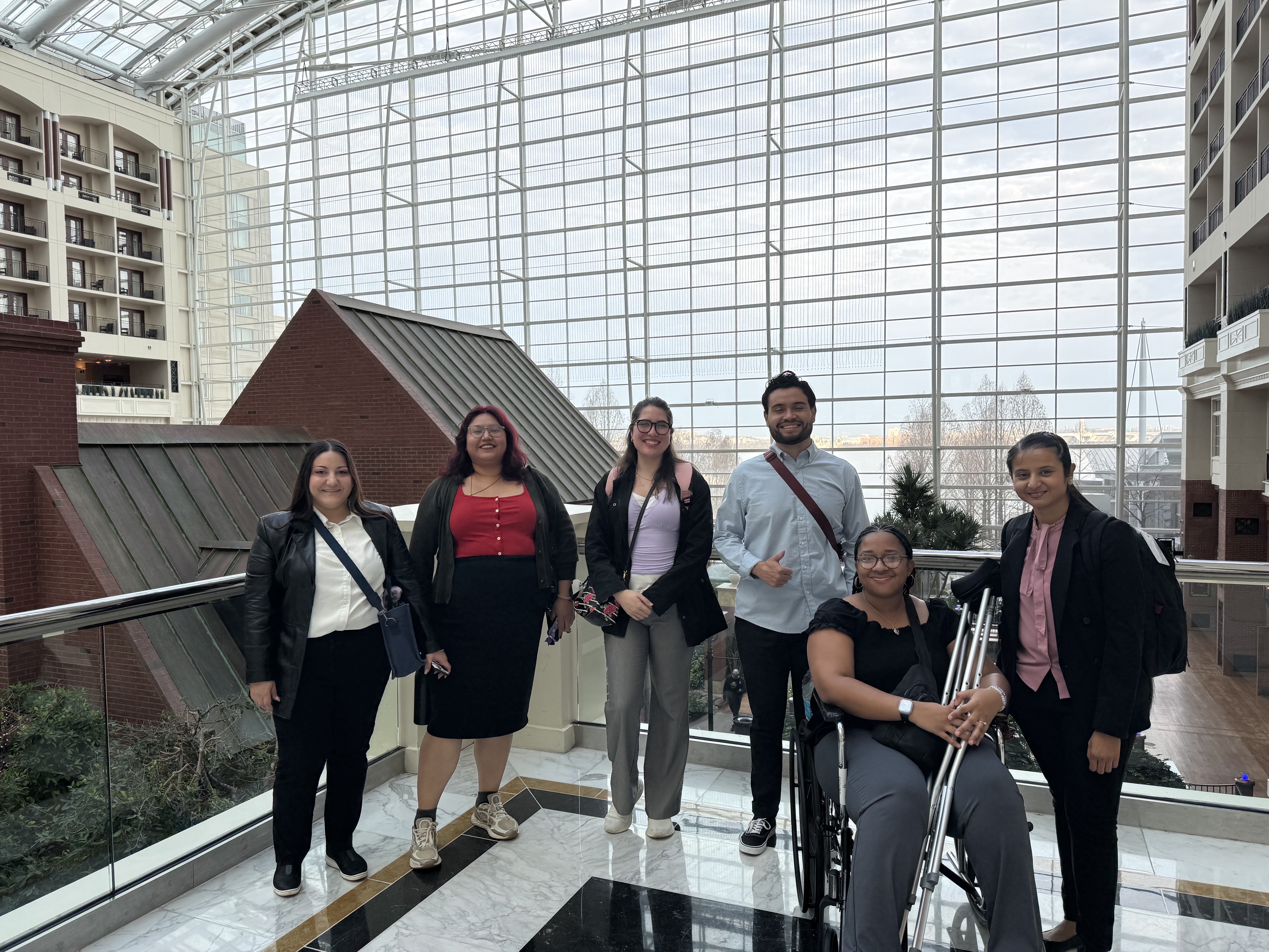 Six professionals pose together in a bright indoor atrium with large glass windows. One person sits in a wheelchair holding crutches, while the others stand beside her, all smiling and dressed in business attire.