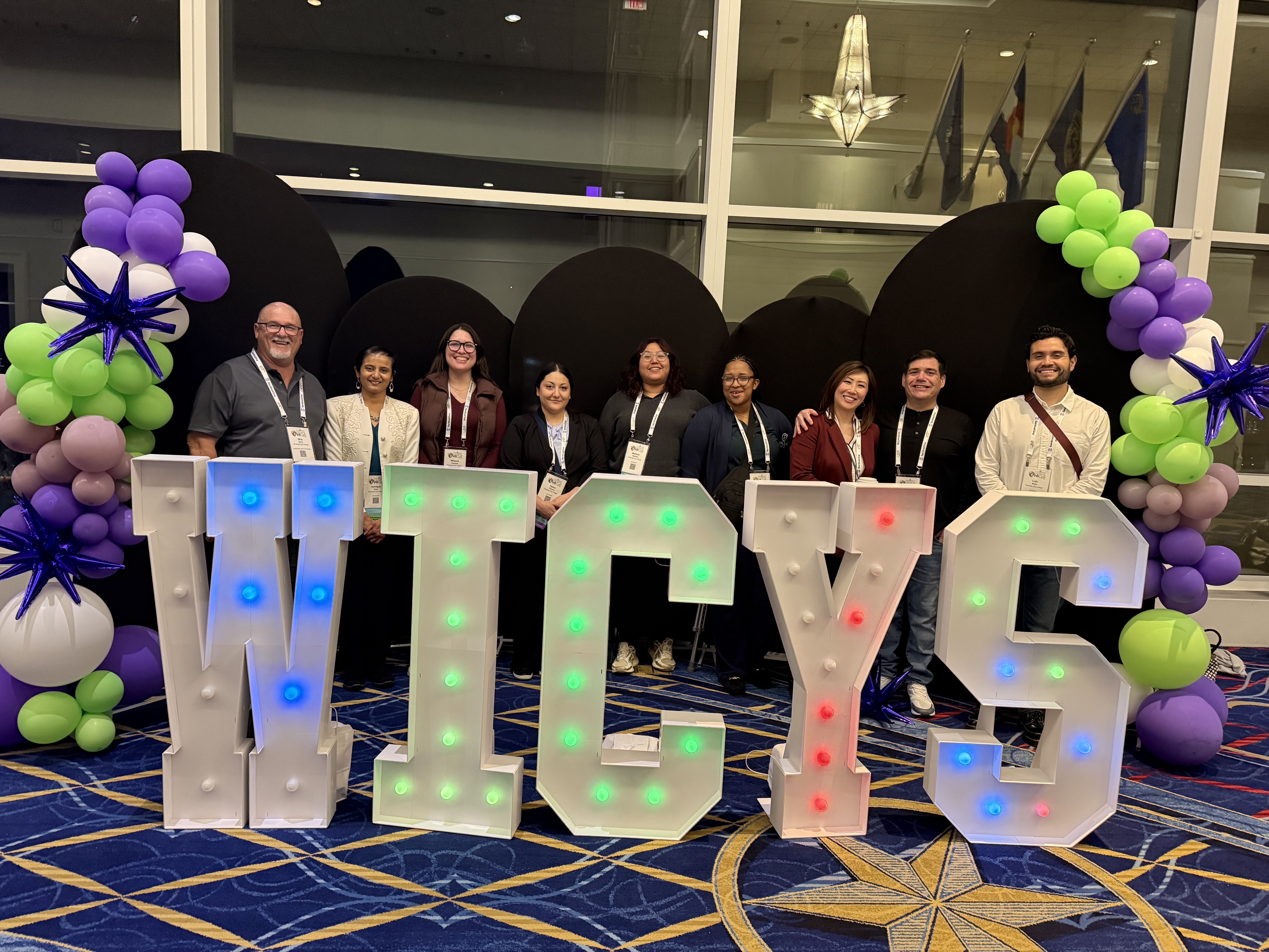 Group of conference attendees pose behind large illuminated letters spelling “WICYS,” surrounded by purple and green balloon decorations in an indoor event space.