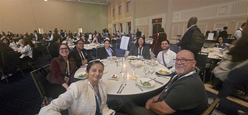 Group of conference attendees sit around a round banquet table during a large conference meal, smiling toward the camera in a crowded ballroom.